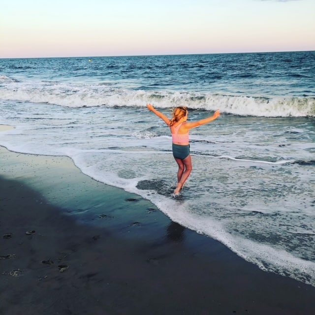 A child with outstretched arms plays in the surf at a beach during sunset, with waves gently crashing onto the shore.