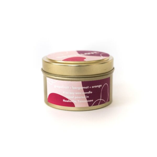 A small gold tin BraveLove Candle labeled "grapefruit + bergamot + orange," with a pink and red abstract design on the lid.