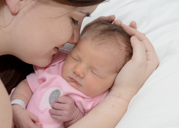 A woman gently holds a sleeping newborn baby in a pink outfit close to her face, lying in bed, sharing a tender moment.