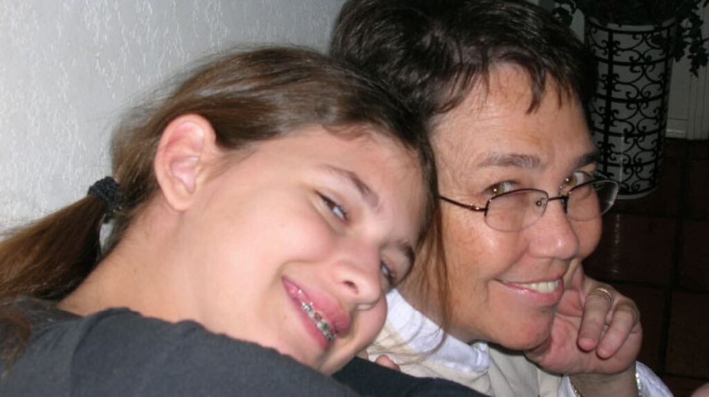 A girl with braces and an adult wearing glasses lean in closely, smiling for a photo indoors.