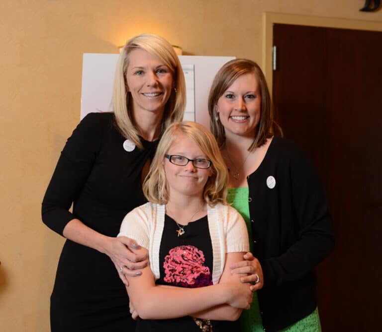 Three smiling people, two adult women and one child, stand together in a group hug with a backdrop in an indoor setting.