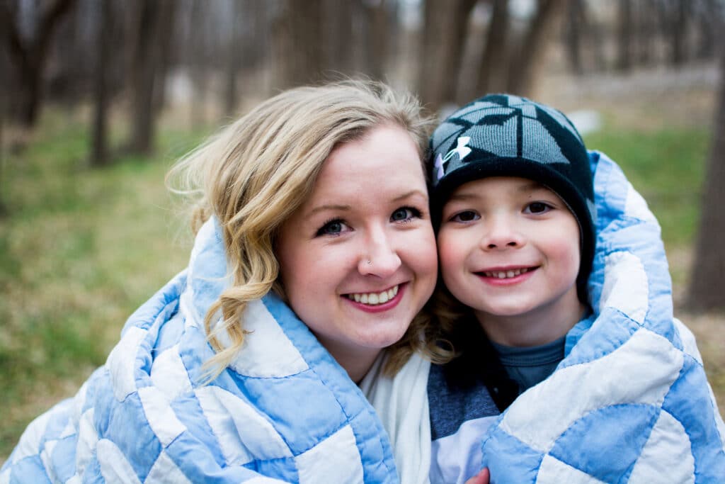 A woman and child, wrapped in a blue and white quilt, smile together outdoors with bare trees in the background.