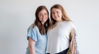 Two women smiling, standing side by side against a plain white background. One wears a blue top and the other, a white top with patterned sleeves.