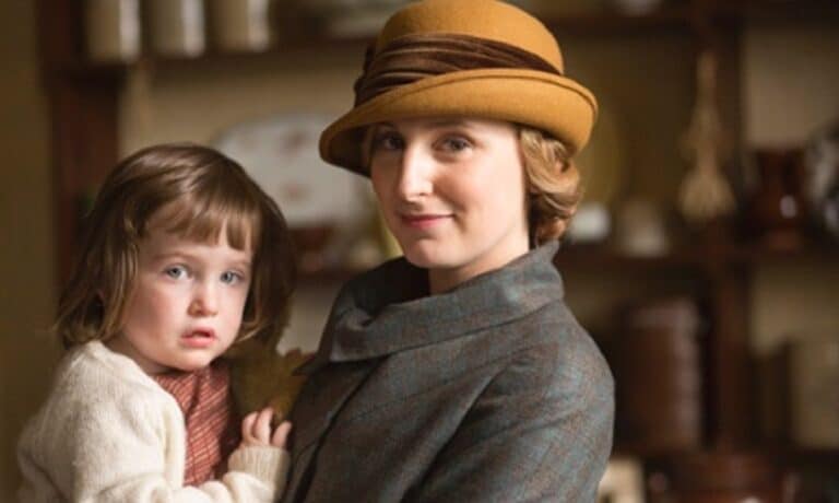 A woman in a brown hat holds a young girl inside a cozy room, both wearing warm clothing and looking at the camera.