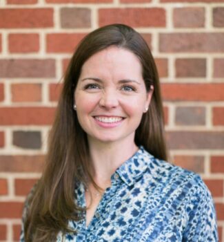 A woman with long brown hair wearing a blue patterned shirt smiles in front of a red brick wall.