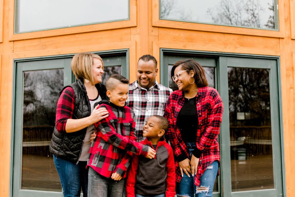 A family of five, dressed in casual attire, gathers in front of a wooden cabin smiling and laughing together.