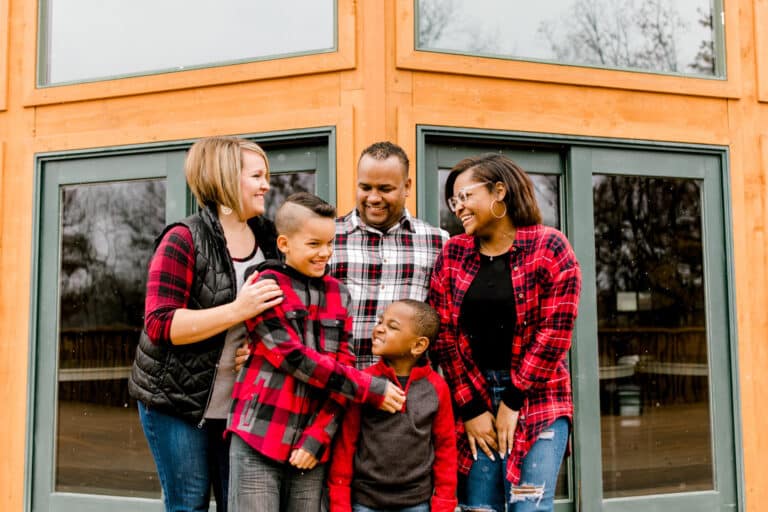 A family of five, dressed in casual attire, gathers in front of a wooden cabin smiling and laughing together.