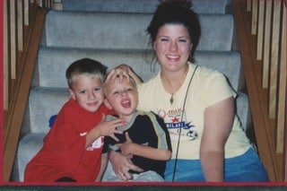 A woman sits on stairs with two young boys; one boy in red hugs the other boy in black, who makes a funny face.