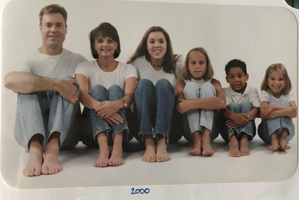 A family of six sitting on the floor against a white background, dressed in white tops and blue jeans, with "2000" written below them.