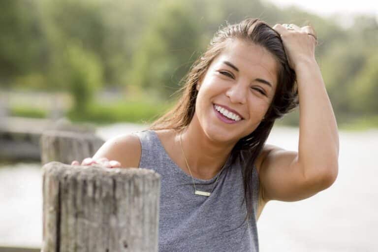 A smiling person with long hair and a sleeveless gray top stands outdoors, touching their hair and leaning on a wooden railing.