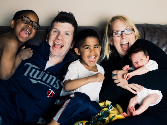 A family of five, including two adults and three children, smiles and laughs while sitting close together on a couch.