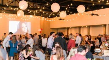 A lively indoor party with people socializing under string lights, balloons, and a large projected image on the wall in the background.