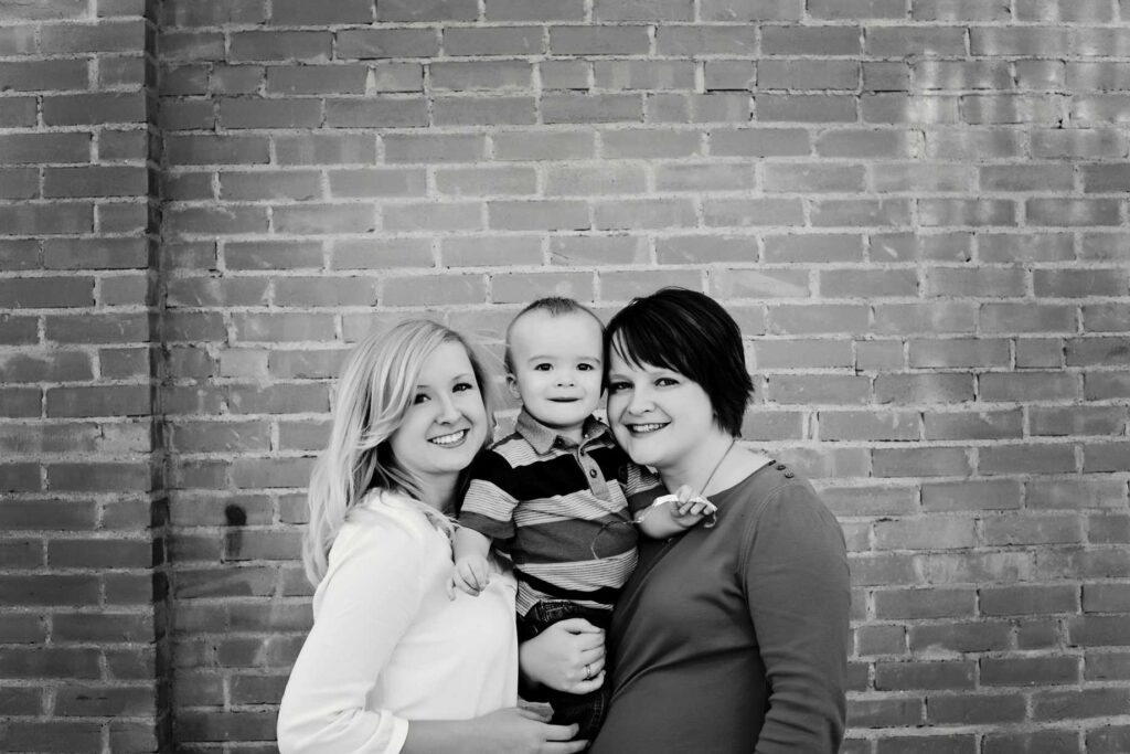 Two women and a child stand in front of a brick wall, smiling at the camera. The child is being held by the woman on the right.