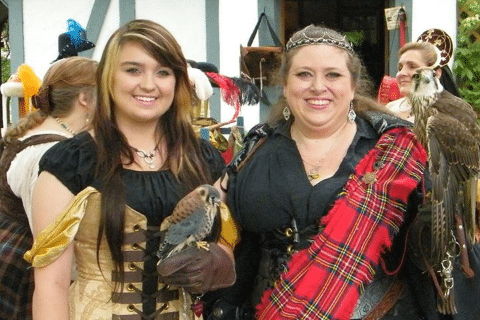 Two women dressed in Renaissance attire hold falcons on their gloved hands while smiling at an outdoor fair.