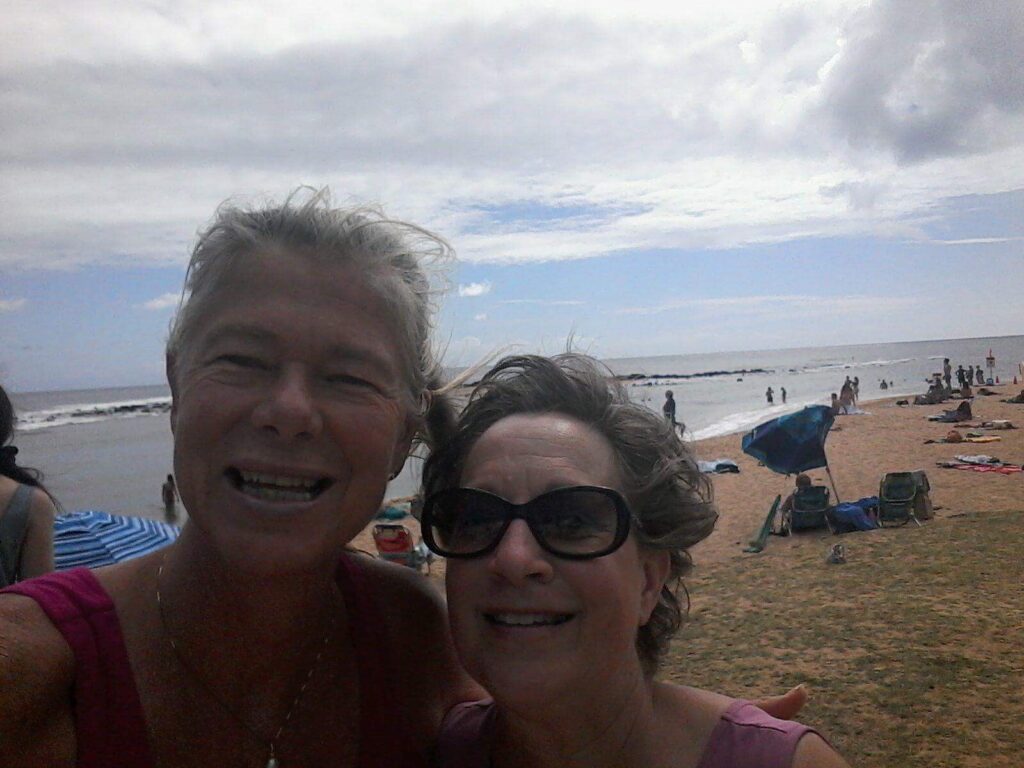 Two people smiling at the beach; one person has short gray hair, and the other person is wearing sunglasses. The ocean and cloudy sky are in the background.