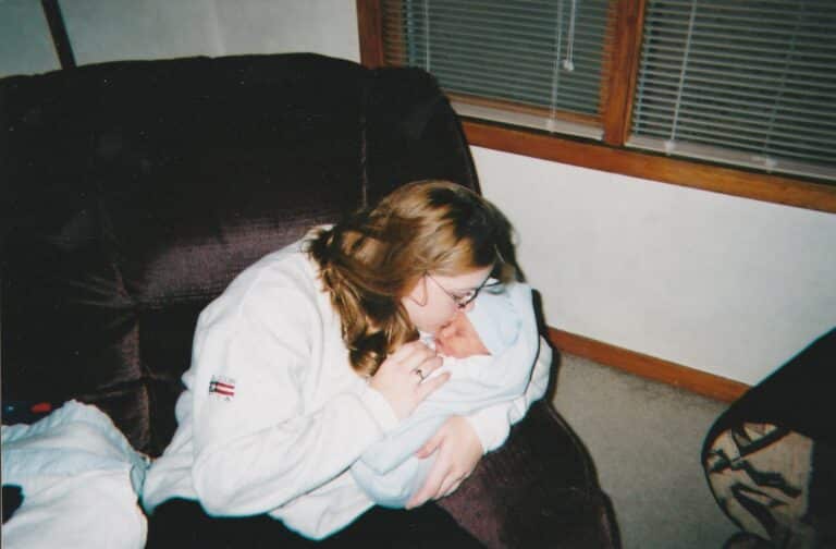 Woman wearing glasses holding and kissing a newborn baby wrapped in a blue blanket while sitting on a dark couch near a window.