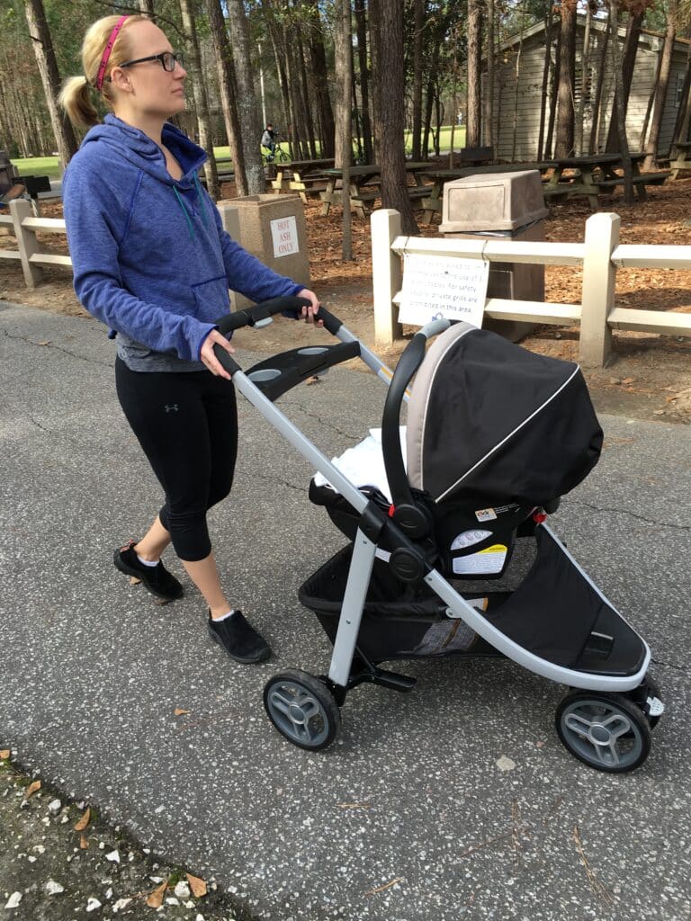 Woman in a blue hoodie and black leggings pushes a stroller on a paved path in a park with trees and a picnic area in the background.