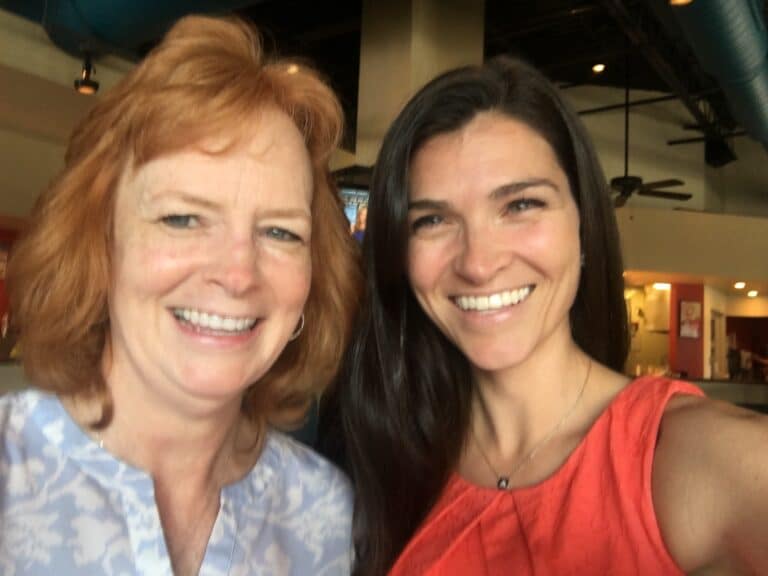 Two women smiling for a selfie indoors; one with red hair wearing a light blue top, the other with dark hair in a coral sleeveless top.