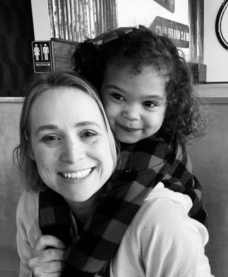 A smiling woman carries a cheerful young girl on her back, both looking at the camera in a cozy indoor setting.