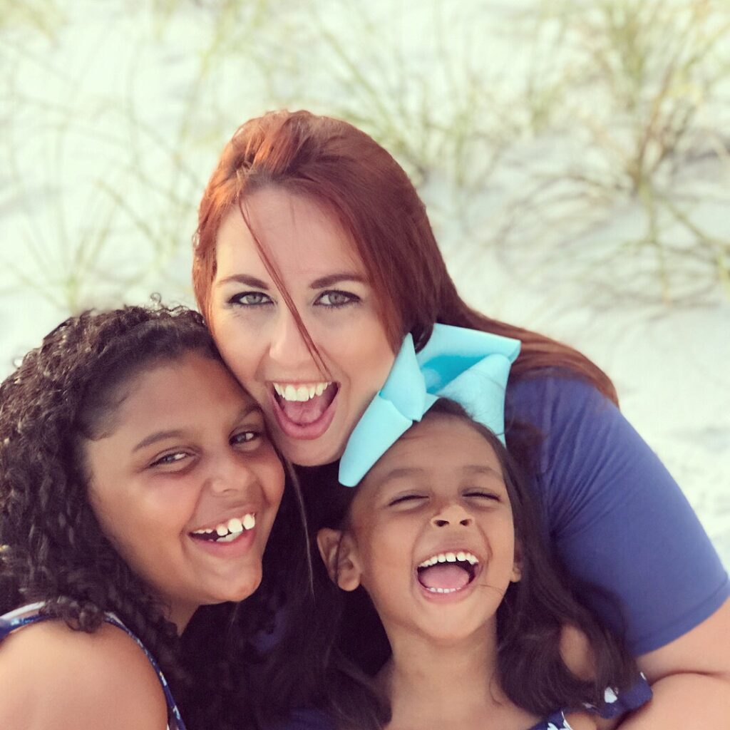 A woman with two young girls, all smiling and laughing, posed closely together outdoors with blurred greenery in the background.