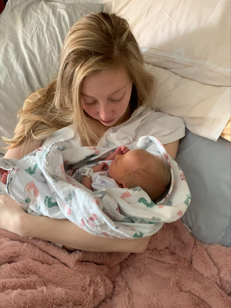 Woman with long hair lies in bed, cradling a newborn wrapped in a blanket covered in multi-colored footprints.