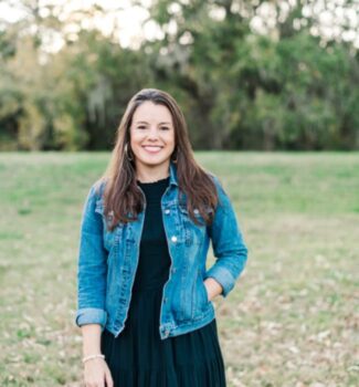 A woman with long hair and a denim jacket stands on grass with trees in the background, smiling at the camera.