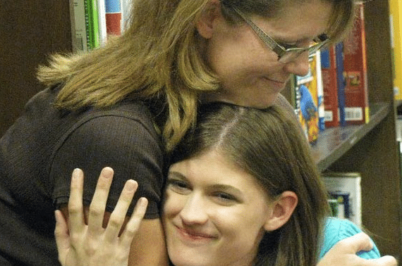 Two people share a warm hug in a library, with books visible in the background. One person smiles brightly while being embraced.