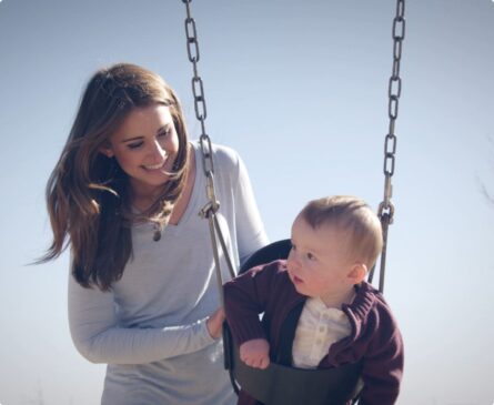 A woman smiles at a baby sitting in a swing under a clear blue sky.