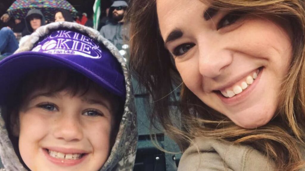 A woman and a child smile at a sporting event; the child is wearing a purple baseball cap and a hoodie. People with umbrellas are visible behind them.