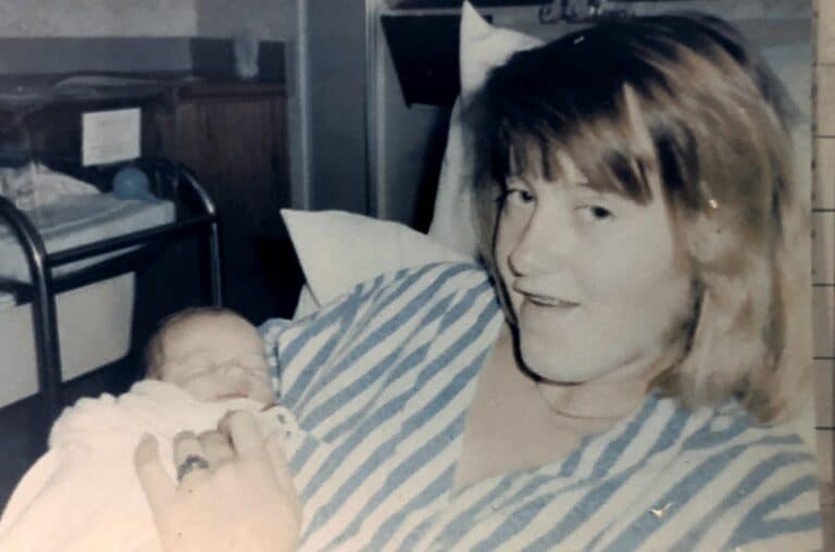 A woman in a striped shirt holds a newborn baby while smiling, hospital crib in the background.