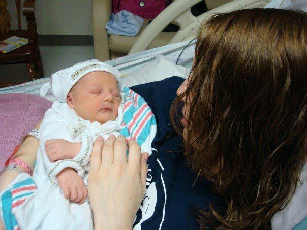 Person with wet hair lying on a hospital bed, holding a sleeping newborn baby wrapped in a blanket, both in white outfits.