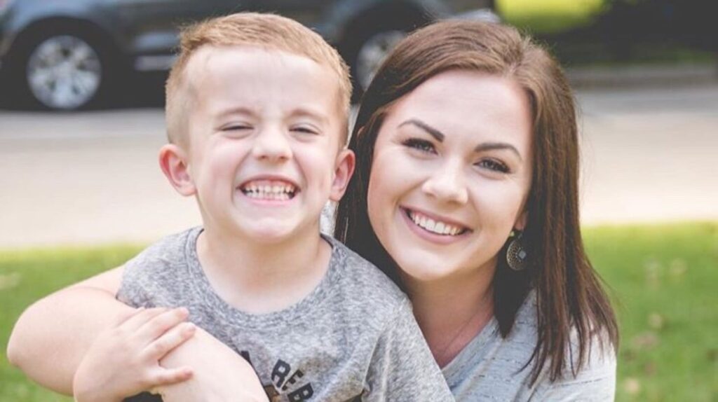 A woman and a young boy, both smiling, pose for a photo outdoors with a car in the background. The woman has an arm around the boy.