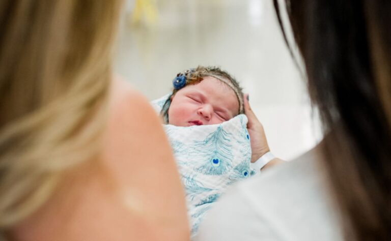 A newborn baby wrapped in a white and blue blanket being held by someone, seen from behind two adults looking on.