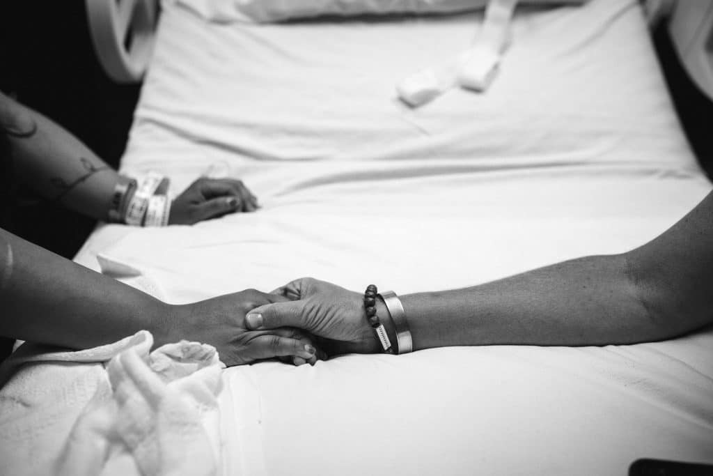 Two people holding hands on a hospital bed, both wearing medical wristbands, in a black and white photograph.