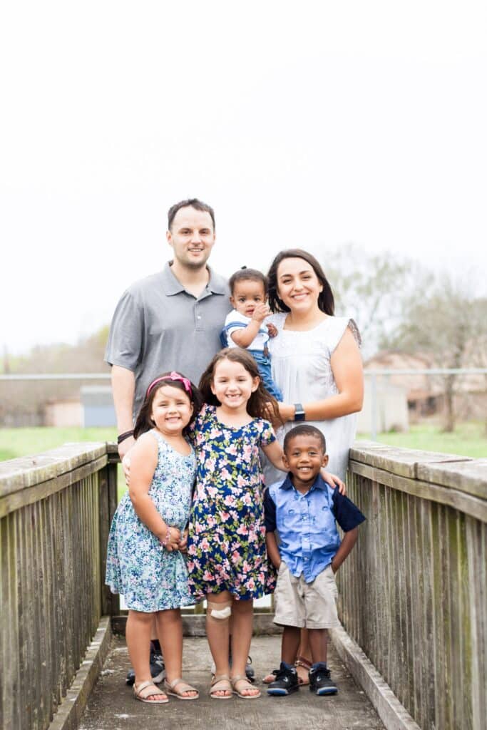 A family of six stands on a wooden bridge. The parents are standing behind four young children, with the mother holding the baby.