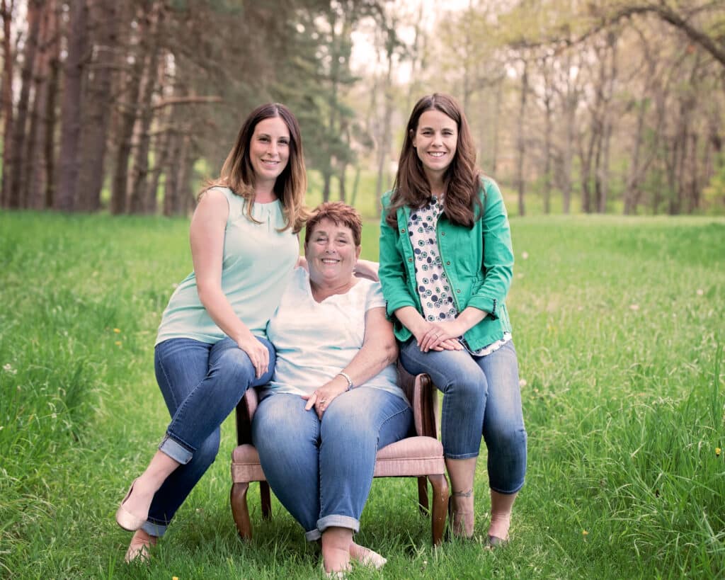 Three women pose for a photo, with one sitting in a chair and the other two standing beside her, in a grassy outdoor setting.