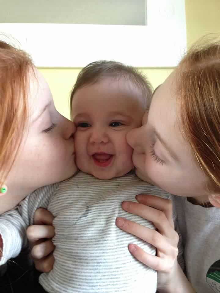 Two people kiss a smiling baby, who is held up close to the camera. The trio share a happy and intimate moment.