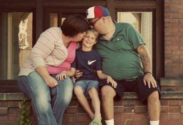 A family of three sits on a window ledge; the parents kiss their smiling child on the head.