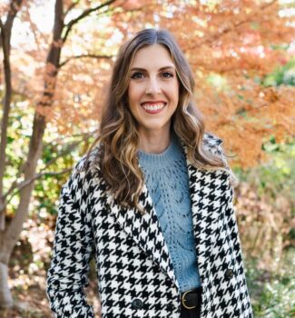 A woman with long brown hair wearing a houndstooth coat and blue sweater smiles in front of a backdrop of autumn trees.