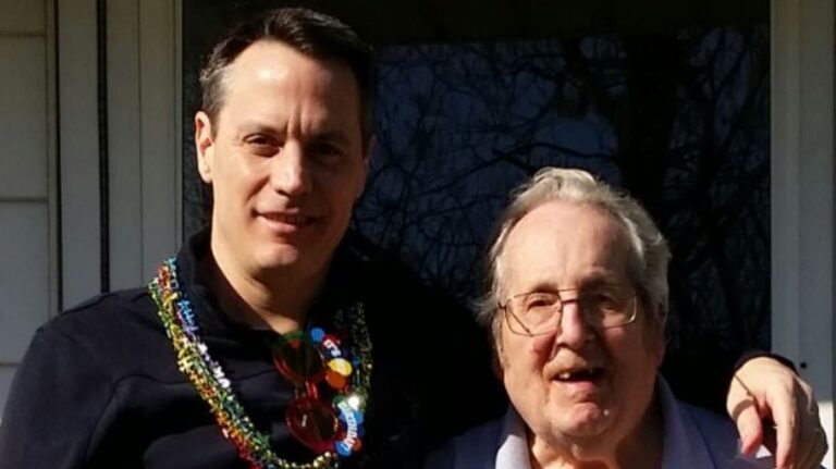 Two men stand outside. The younger man wears colorful beads and sunglasses around his neck, the older man smiles and wears glasses.