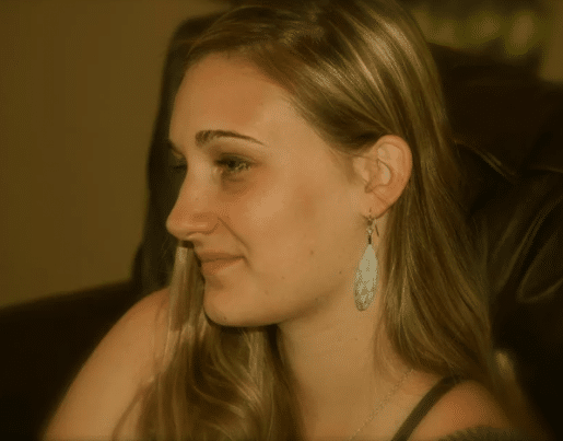 A woman with long hair and light earrings sits, slightly smiling, and looks off to the side against a blurred brown background.