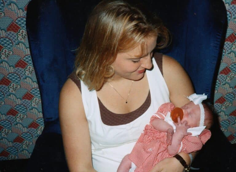 A woman with blonde hair smiles down at a baby in a red and white dress, the baby has a pacifier in its mouth and a white bow on their head.