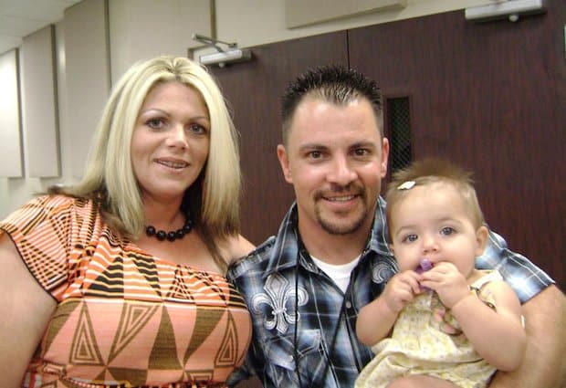 A woman and man with a baby in the man's arms, all smiling at the camera indoors near a door with a window.