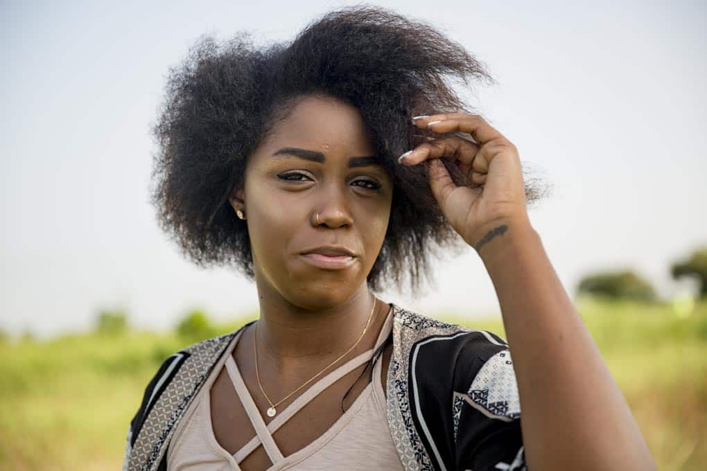 A person with an afro hairstyle is standing outdoors, tilting their head slightly and touching their hair, with a green field in the background.