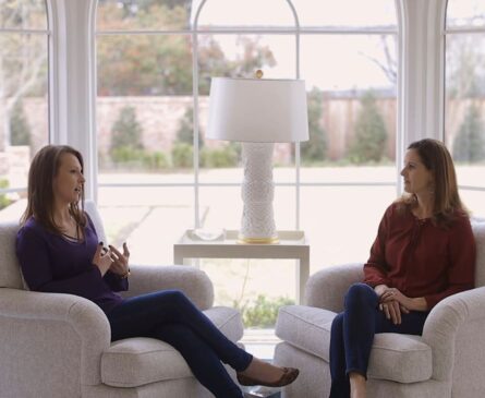 Two women sit in armchairs facing each other by an arched window, engaging in conversation; a lamp sits on a table between them.