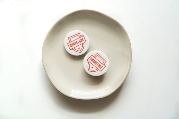 Two BraveLove Popsockets, featuring "BRAVELOVE" printed in red on a white backdrop, arranged on a white plate against a seamless white background.