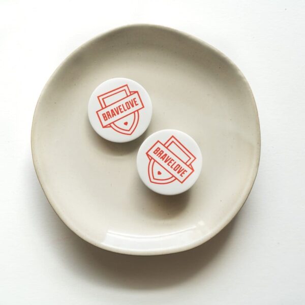 Two BraveLove Popsockets, featuring "BRAVELOVE" printed in red on a white backdrop, arranged on a white plate against a seamless white background.