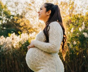 A pregnant woman in a white sweater stands in a sunlit field, cradling her belly and smiling while looking into the distance.