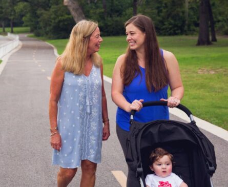 Two women walking on a park path, one pushing a stroller with a baby inside. They are smiling and engaged in a cheerful conversation.