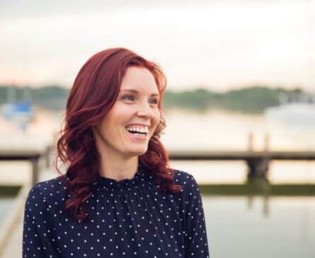 A woman with red hair wearing a black polka dot blouse is smiling, standing in front of a blurry waterfront background.